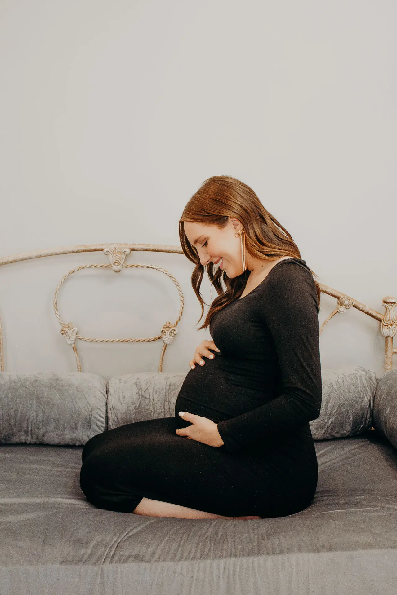 Pregnant woman in a long black dress on her knees on a bed holding her baby bump